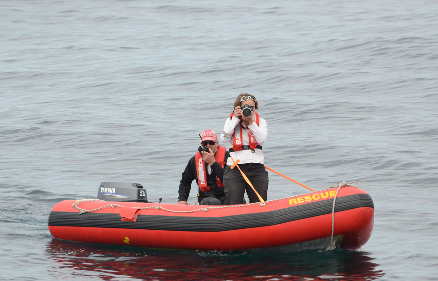 Two people in life jackets are on a small red inflatable rescue boat in open water, one using a camera and the other speaking into a radio.