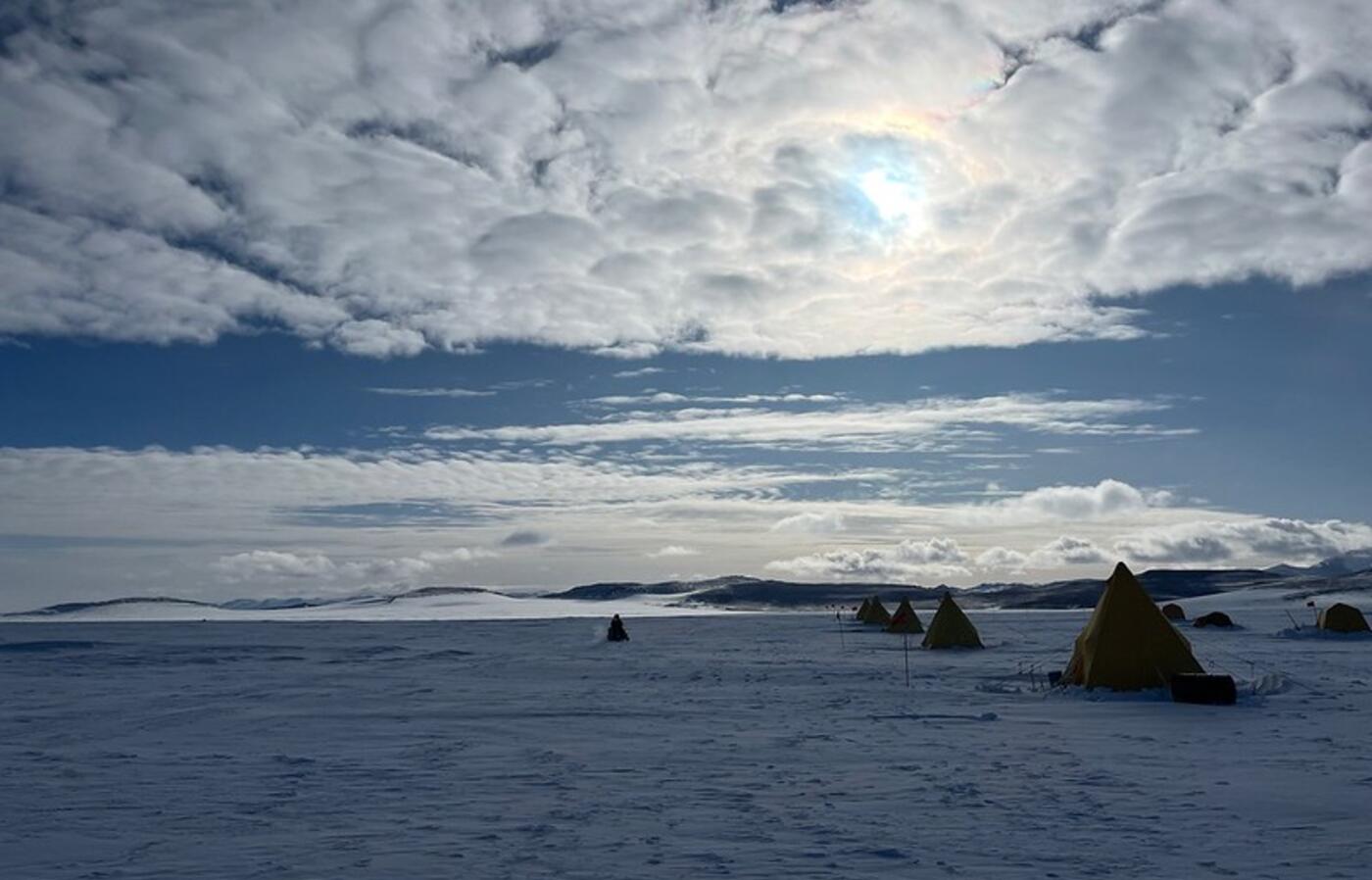 Snowy Antarctic landscape dotted with tents from a research field camp in the Allan Hills, with clouds, sun and blue skies. 
