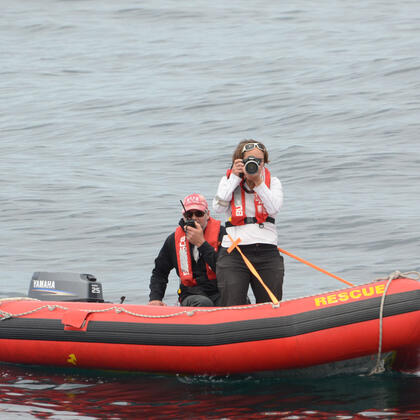 Two people in life jackets are on a small red inflatable rescue boat in open water, one using a camera and the other speaking into a radio.