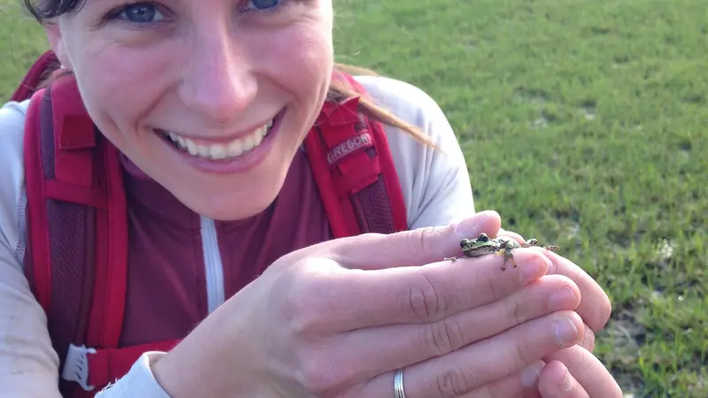 A woman smiles as she holds a tiny green frog in her hands.