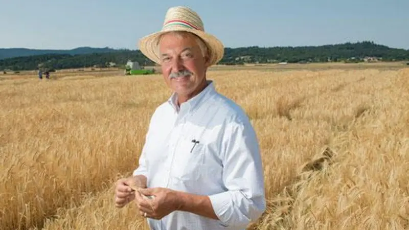 A man wearing a hat smiles as he stands in a yellow barley field. Mountains are in the background.