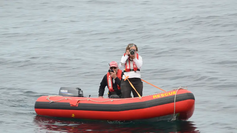 Two people in life jackets are on a small red inflatable rescue boat in open water, one using a camera and the other speaking into a radio.