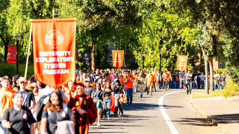 Students in orange, carrying banners, march along a road, surrounded by green trees.