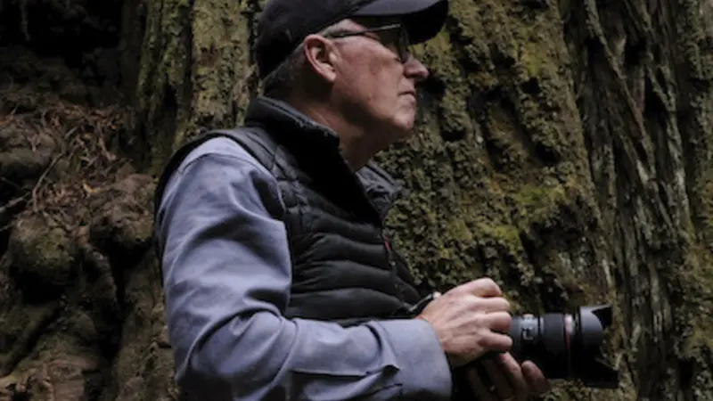 Photographer Chris Johns appears in a black vest and baseball cap, facing away from the camera to the right amid a stand of big trees