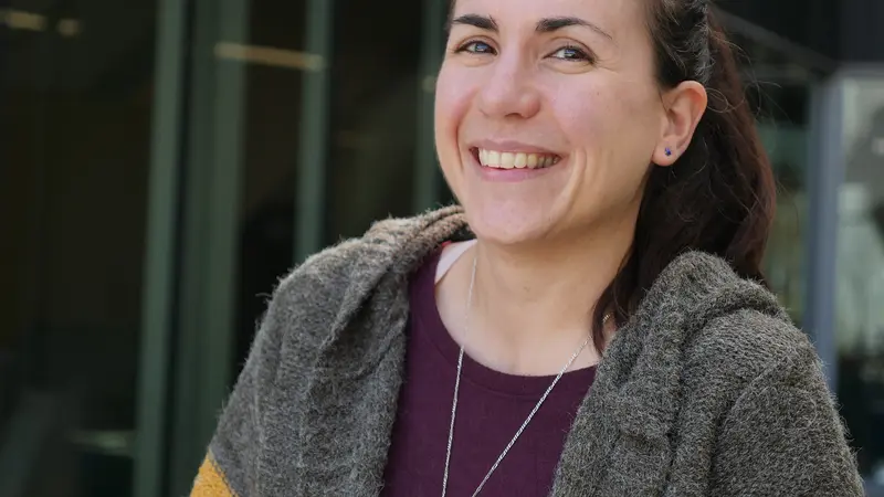 A woman with brown hair is wearing a gray sweater and smiling while posing for a photo.