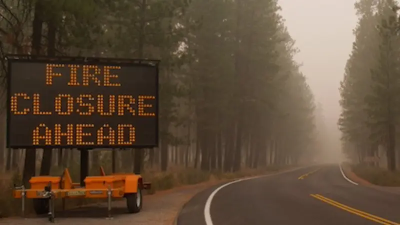 Image shows a large electronic road sign to the left of a highway announcing "Fire closure ahead." The road winds through a smoke-filled forest.