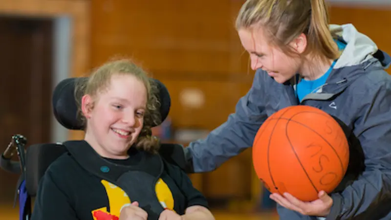 Image shows a volunteer holding a basketball and smiling with a student participant in a wheelchair. Both are in a gymnasium, participating in OSU's IMPACT program.
