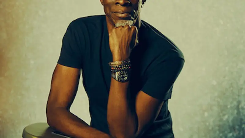 Musician Keb' Mo' looks directly at the camera, sitting up with his guitar atop his lap and resting his chin on his fist. He wears a tan fedora-style hate and a dark t-shirt and pants. He looks very cool.