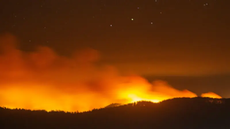 Image of red-orange glow of wildfire and smoke creeping over a darkened hillside at night