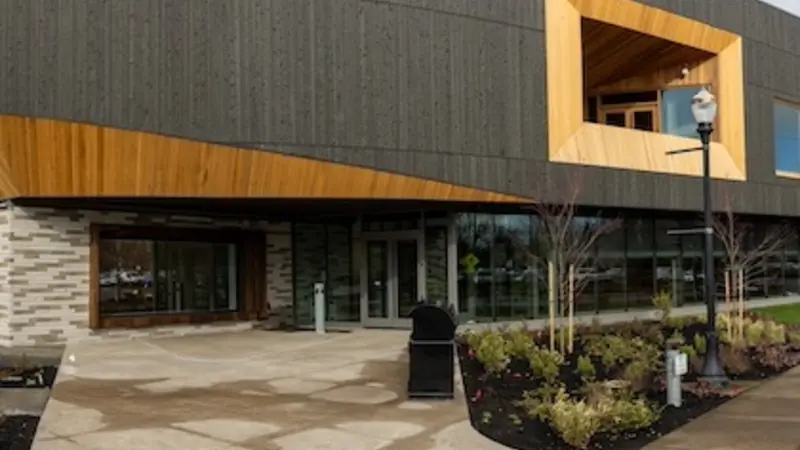 Wide-angle shot of the main entrance of PRAx, a new building on OSU's campus with a dark gray overhanging second story, large inset windows trimmed in blond wood and a wall of windows showing the first-floor lobby. 