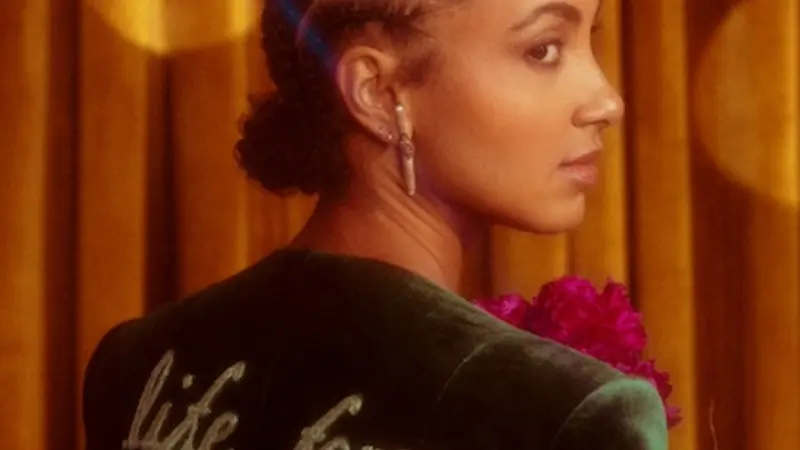 Image shows jazz artist esperanza spalding in front of a copper-colored curtain; she's wearing a green velvet jacket with "life force" written across the back, and she's looking at the camera over her right shoulder. 