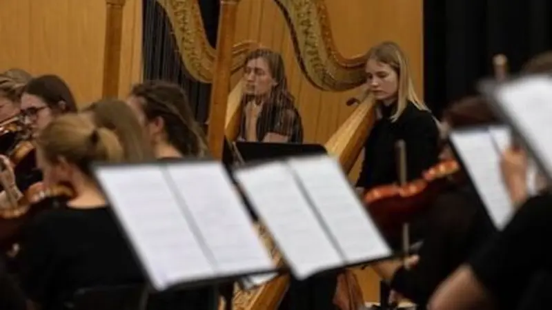 Image of women playing instruments in a symphonic orchestra, with the camera focusing on a young blonde woman sitting at a large harp in the center.