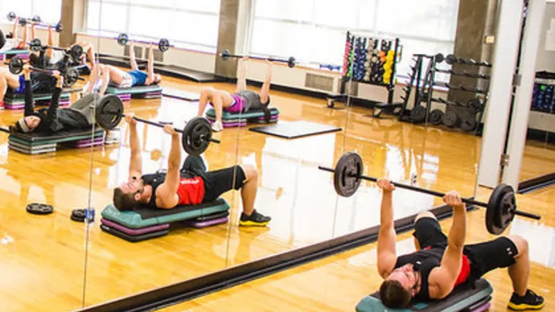An instructor leads students in a Body Pump class at OSU; students are arrayed about a fitness room in front of a wall of mirrors, lying back on stacks of risers and raising barbells over their heads.
