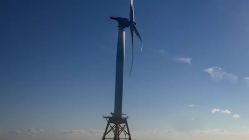 Image shows a series of 5 massive offshore wind turbines in open ocean under a glaring sun; the first turbine is close by in the foreground and the others look progressively smaller as they are farther away.