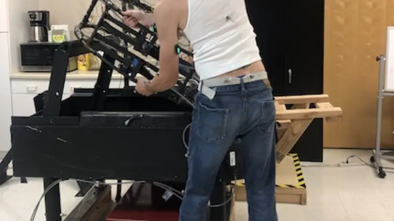 Image shows a man in a white tank top and blue jeans from behind, standing in front of a table holding an empty metal crab pot, with electrodes connected to different points of his upper body to record muscle activiity.
