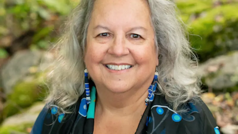 Headshot of author Robin Wall Kimmerer, a smiling lady with shoulder-length gray hair, wearing blue and white beaded earrings and a blue, black and turquoise shawl.