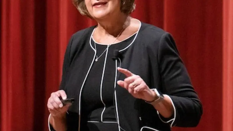 Ruth Watkins speaks onstage in front of the orange curtain at OSU's University Day, wearing a black dress with white piping and a matching blazer.