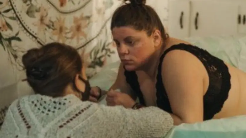 A dark-haired woman in labor leans against the edge of a water-birth tub while a birthing coach crouches next to her. 