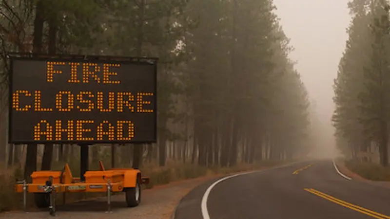 In a smoky forest, a large road sign on the left of a two-lane road says "Fire Closures Ahead." Image was taken during the 2020 wildfires in Oregon. 
