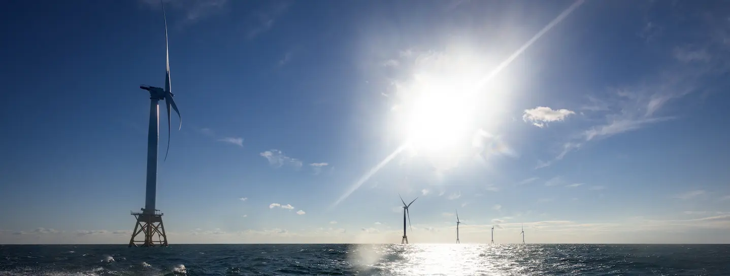 Image shows a series of 5 massive offshore wind turbines in open ocean under a glaring sun; the first turbine is close by in the foreground and the others look progressively smaller as they are farther away.
