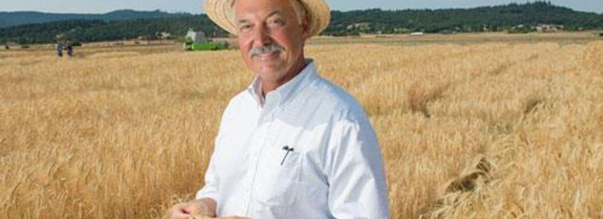 A man wearing a hat smiles as he stands in a yellow barley field. Mountains are in the background.