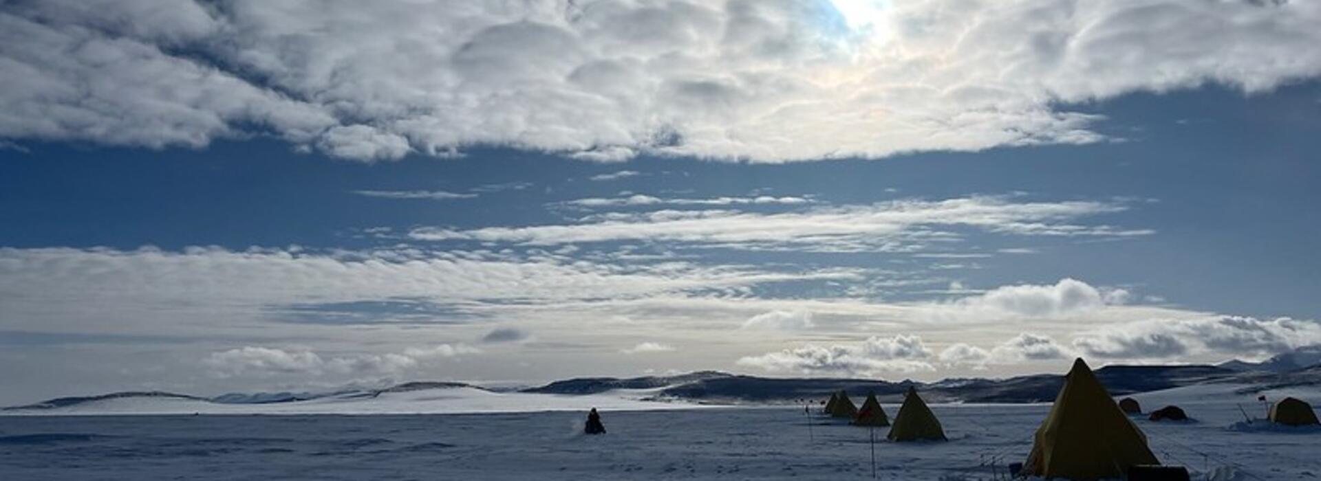 Snowy Antarctic landscape dotted with tents from a research field camp in the Allan Hills, with clouds, sun and blue skies. 