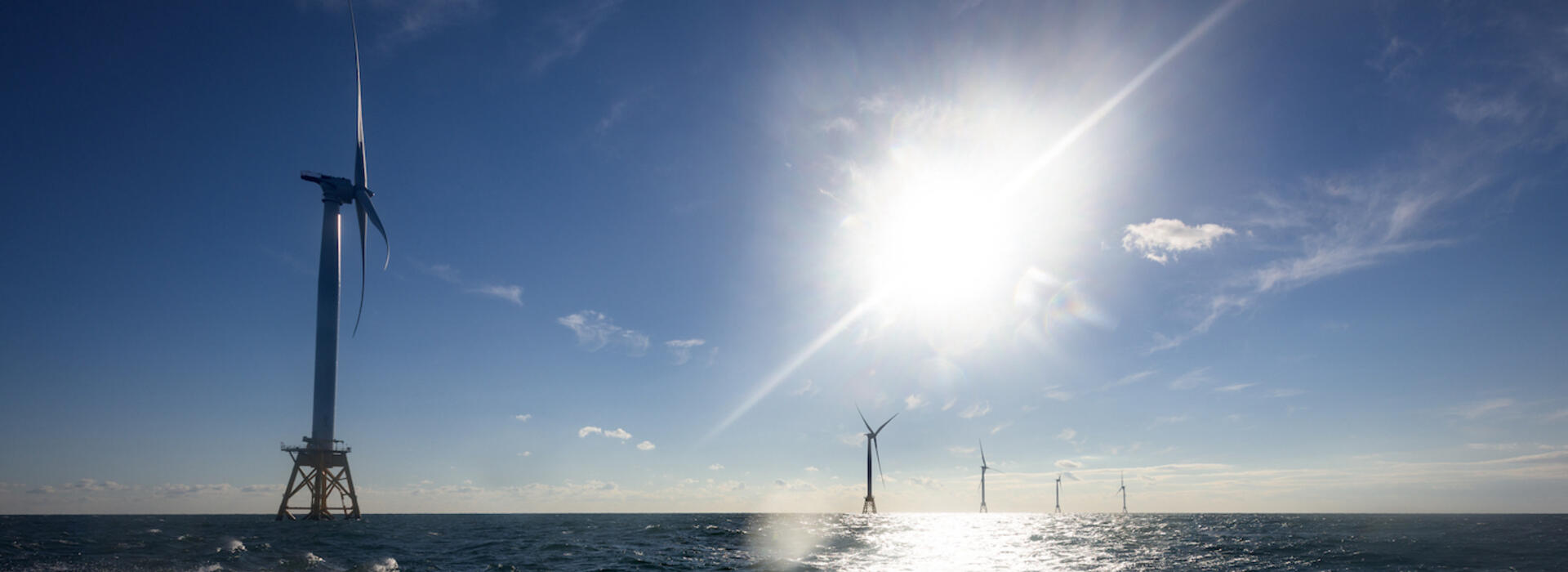 Image shows a series of 5 massive offshore wind turbines in open ocean under a glaring sun; the first turbine is close by in the foreground and the others look progressively smaller as they are farther away.