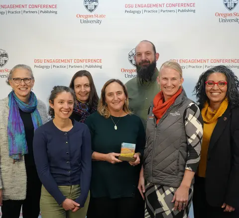 A group of seven people smile together in front of an Oregon State University backdrop, with one person holding an award.