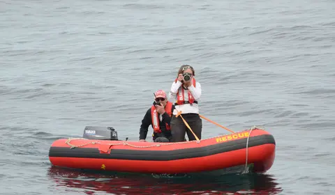 Two people in life jackets are on a small red inflatable rescue boat in open water, one using a camera and the other speaking into a radio.