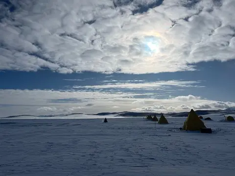 Snowy Antarctic landscape dotted with tents from a research field camp in the Allan Hills, with clouds, sun and blue skies. 